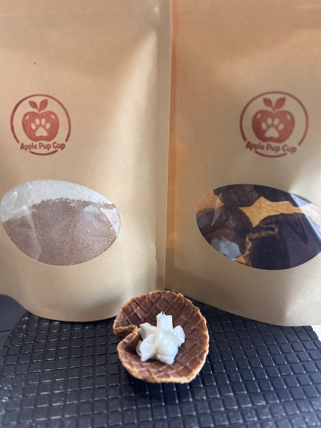 Two kraft Apple Pup Cup bags side by side — one holding the dry batch-kit mix, one holding baked pup cups — with a finished cup topped with whipped cream in front.
